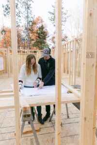 Workers review house plans beside a framed home with foundation work and construction materials nearby.