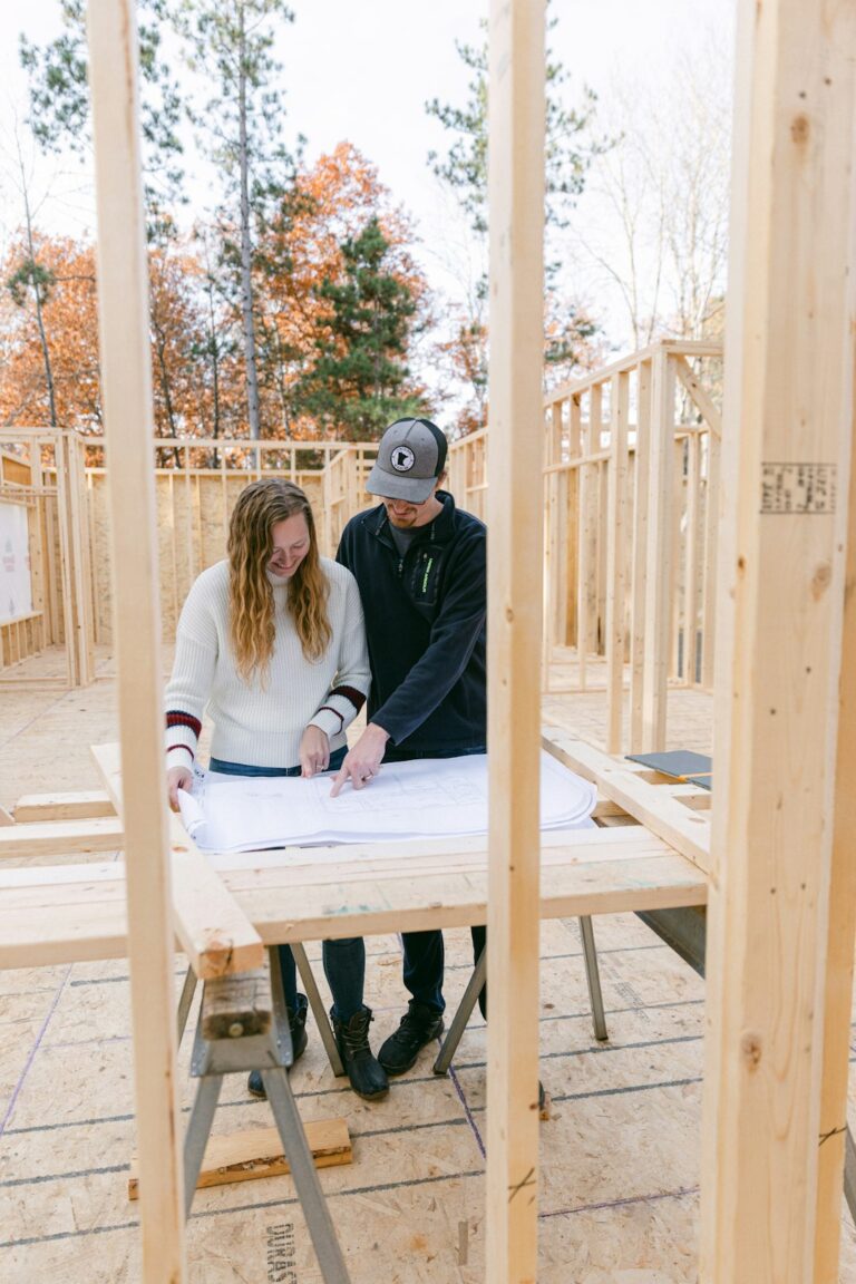 Workers review house plans beside a framed home with foundation work and construction materials nearby.